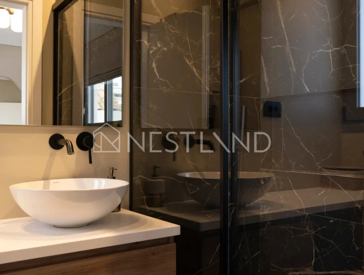 Modern bathroom with black marble walls, glass shower, and white vessel sink on wooden vanity.