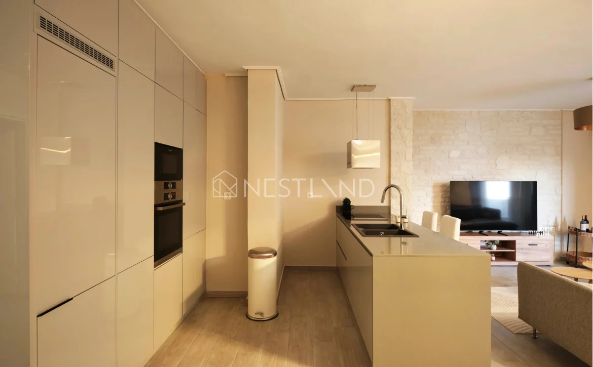 Modern kitchen with white cabinets, built-in appliances, and an island, adjacent to a living area with a stone wall.