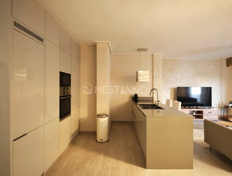 Modern kitchen with white cabinets, built-in appliances, and an island, adjacent to a living area with a stone wall.