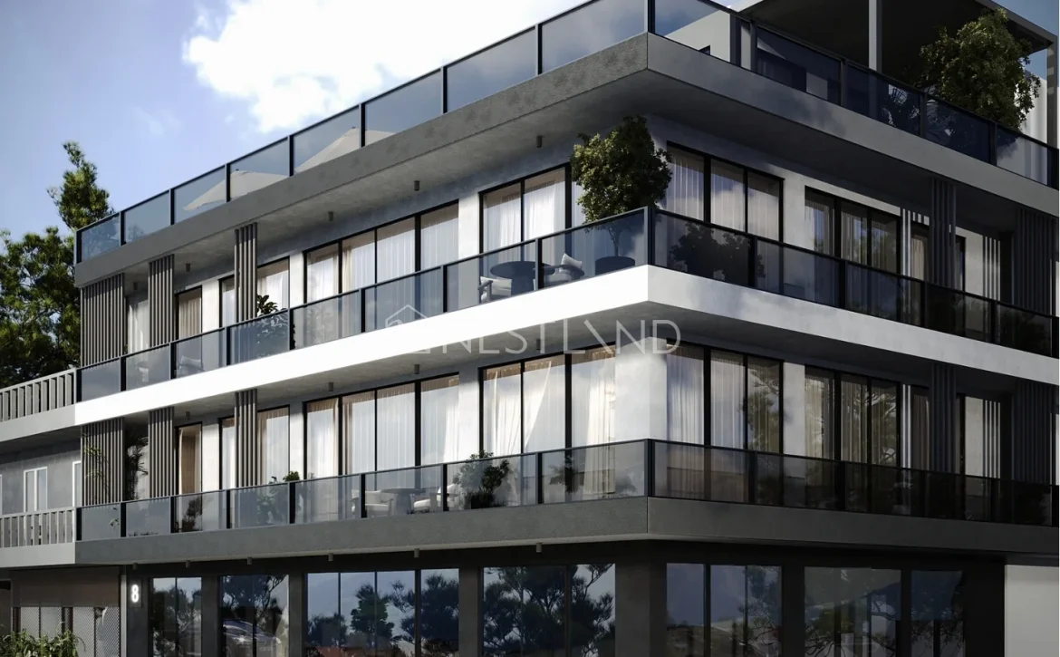 Modern multi-story building with large glass balconies and plenty of natural light, surrounded by greenery and a clear blue sky.