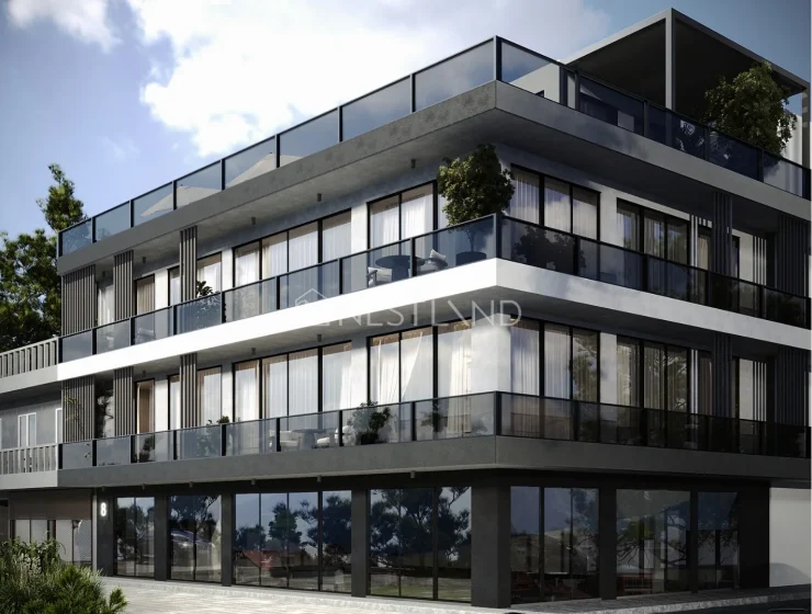 Modern multi-story building with large glass balconies and plenty of natural light, surrounded by greenery and a clear blue sky.