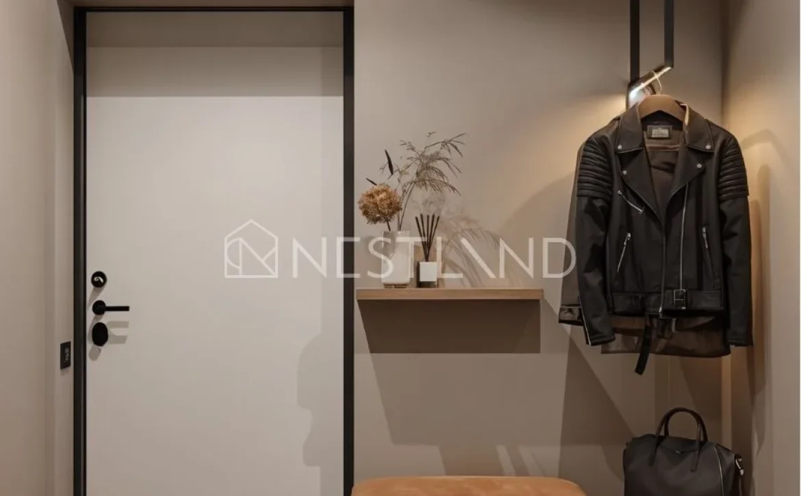 Modern entryway featuring a white door, leather jacket on a hanger, stylish bench, decorative vase, and shoes on a slatted shelf.